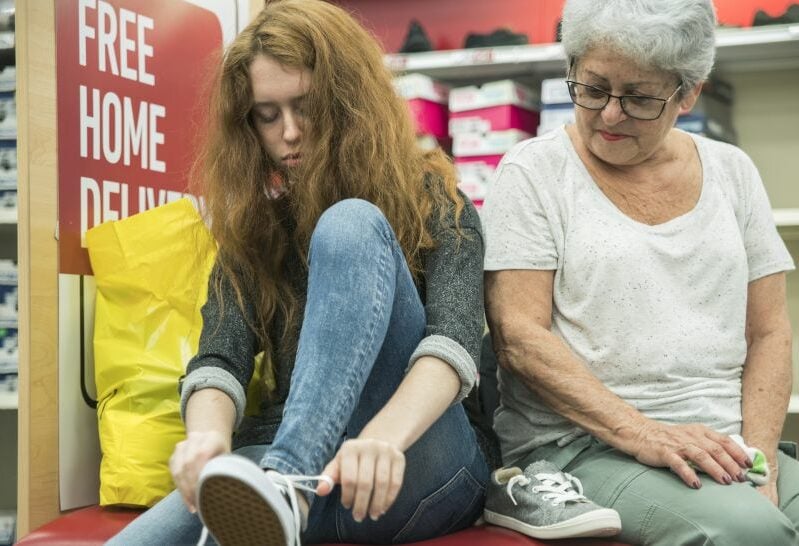 A girl tries on shoes in a shop as an older woman looks on.